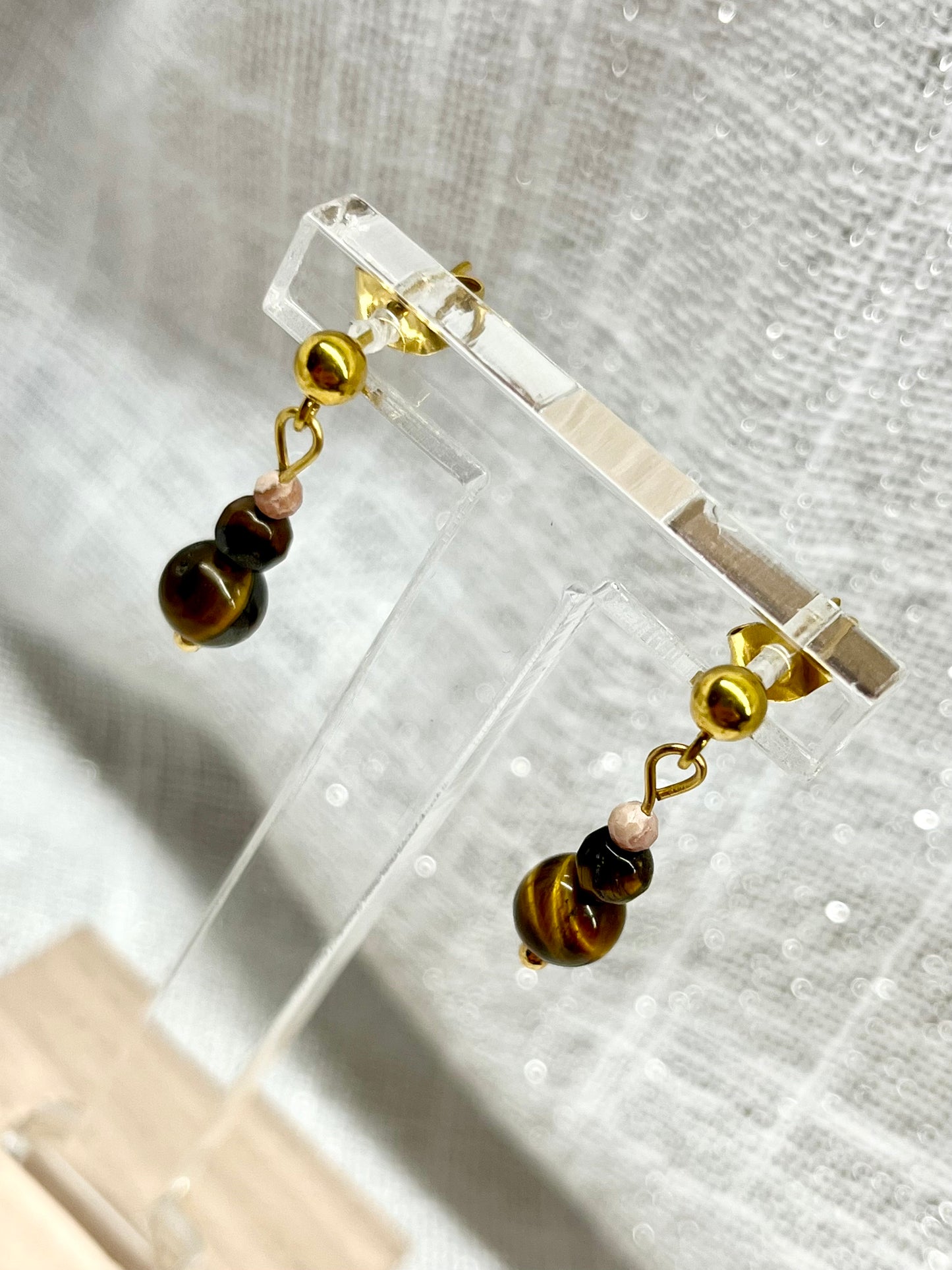 Boucles d'oreille EMMA, aevc pierre d'Oeil de Tigre et de Rhodochrosite, exposées sur un fond en lin.