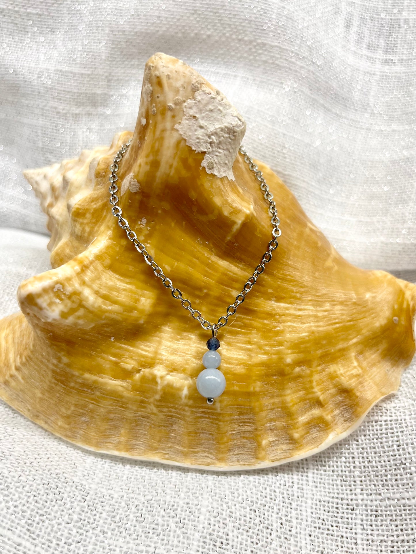 Collier EMMA, avec pierres d'Angélite et de Saphir, exposée sur un coquillage.