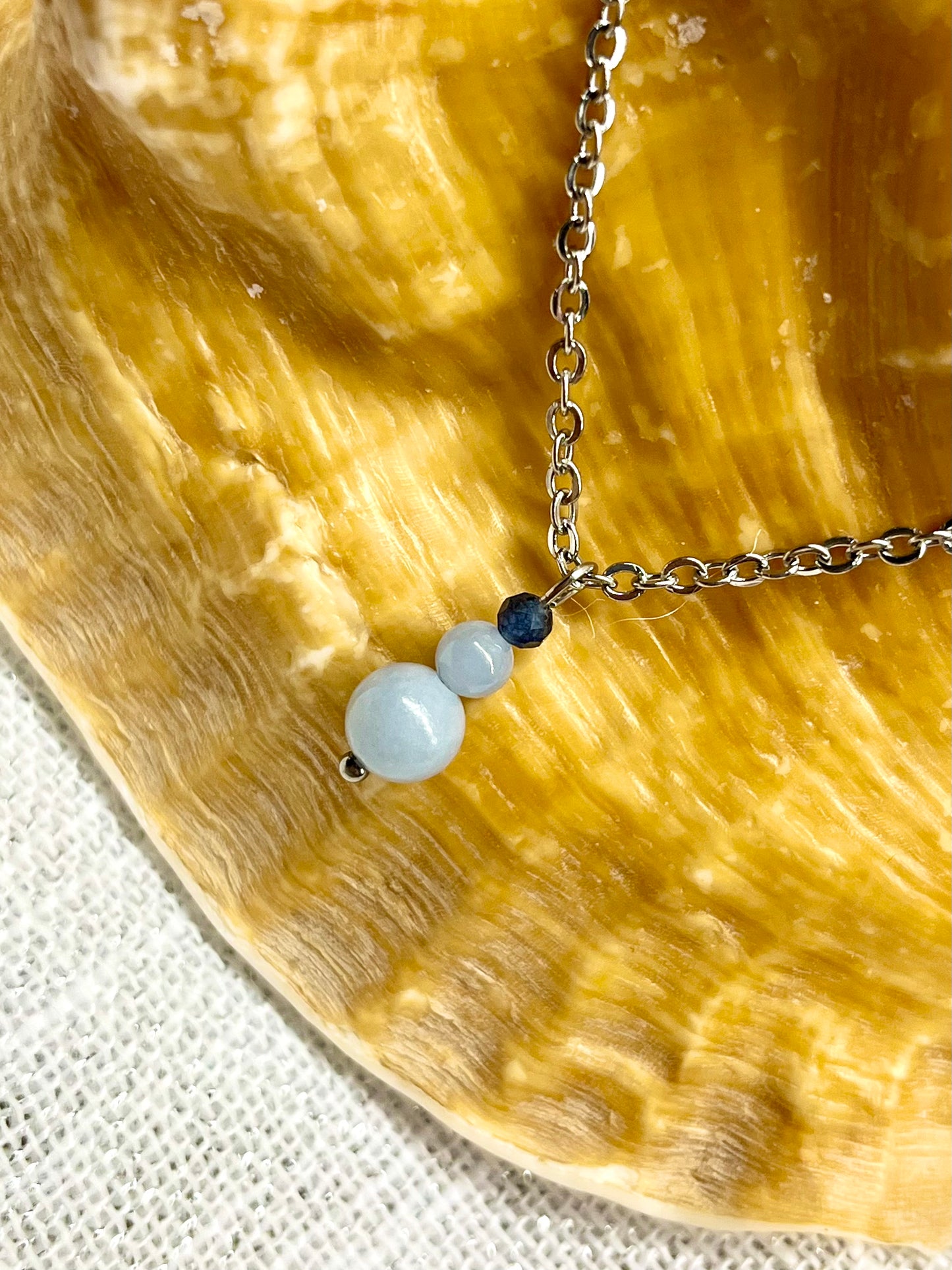 Collier EMMA, avec pierres d'Angélite et de Saphir, exposé sur un coquillage.