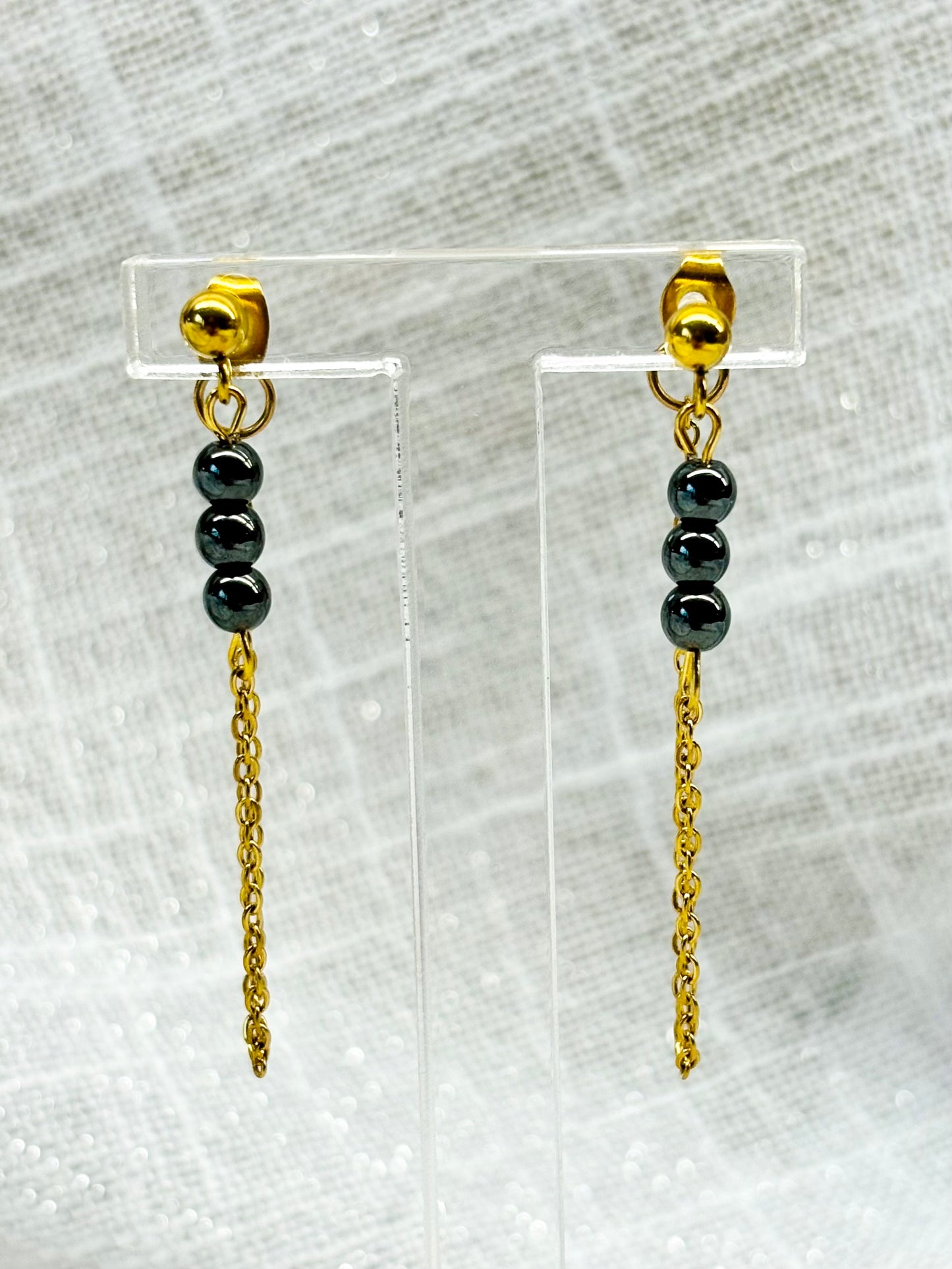 Boucles d'oreille MALIA, de couleur dorée avec pierres d'hématite, exposées sur un fond en lin.