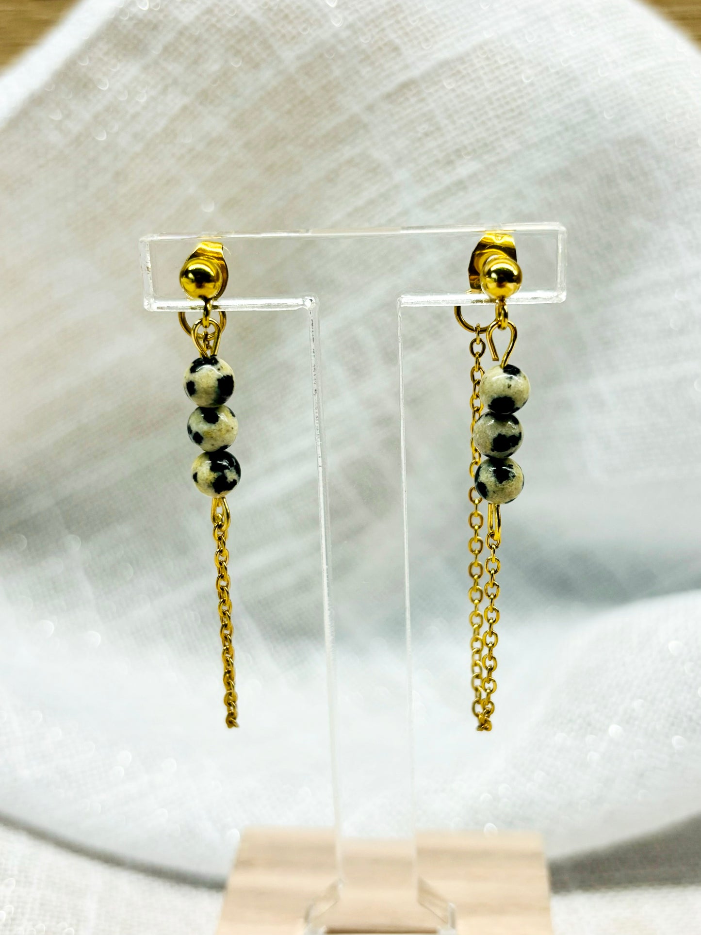 Boucles d'oreille MALIA, de couleur dorée avec pierre de Jaspe Dalmatien, exposées devant un fond en lin.
