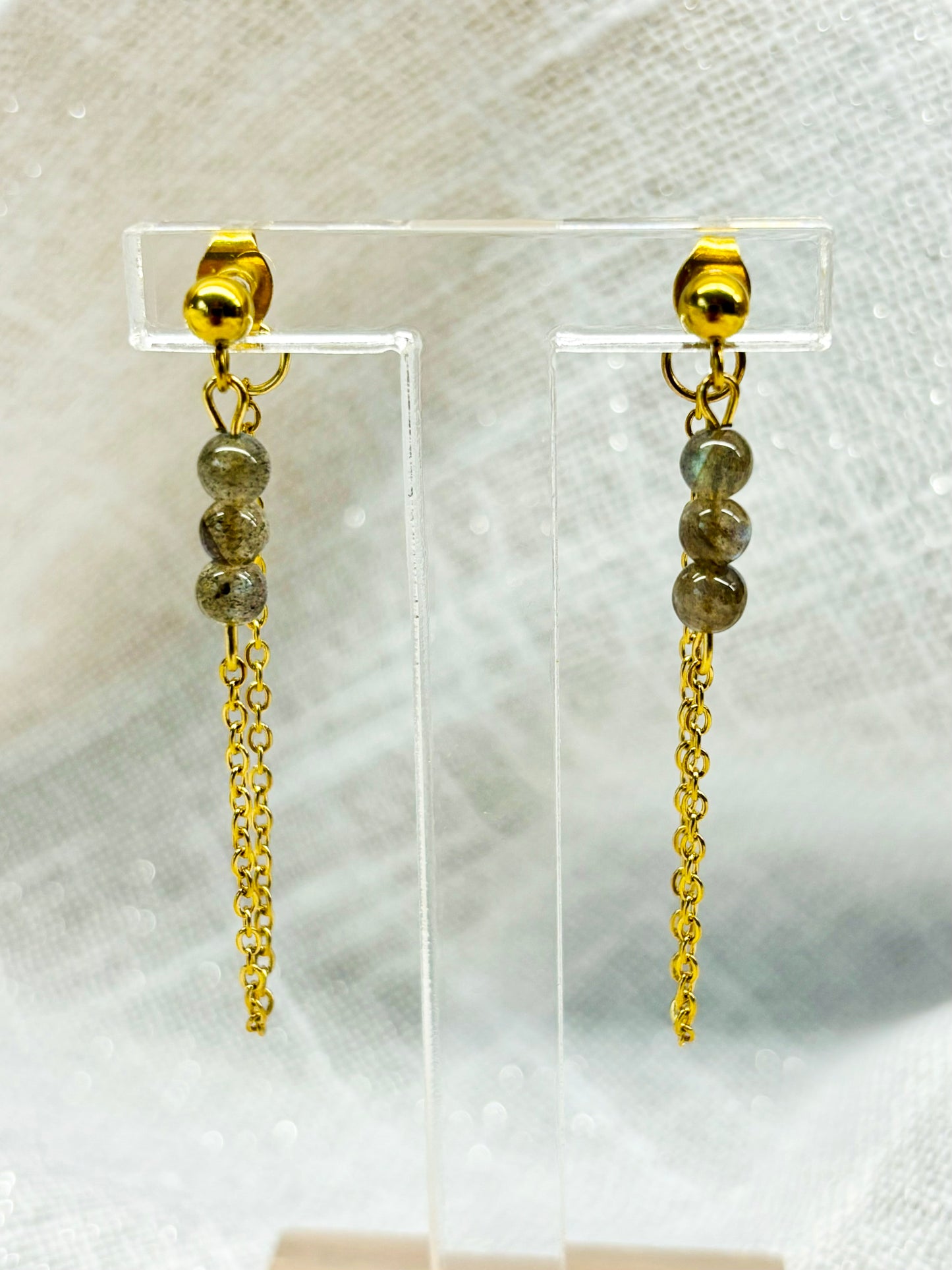 Boucles d'oreille MALIA, de couleur dorée avec pierres de Labradorite, exposées sur un fond en lin.