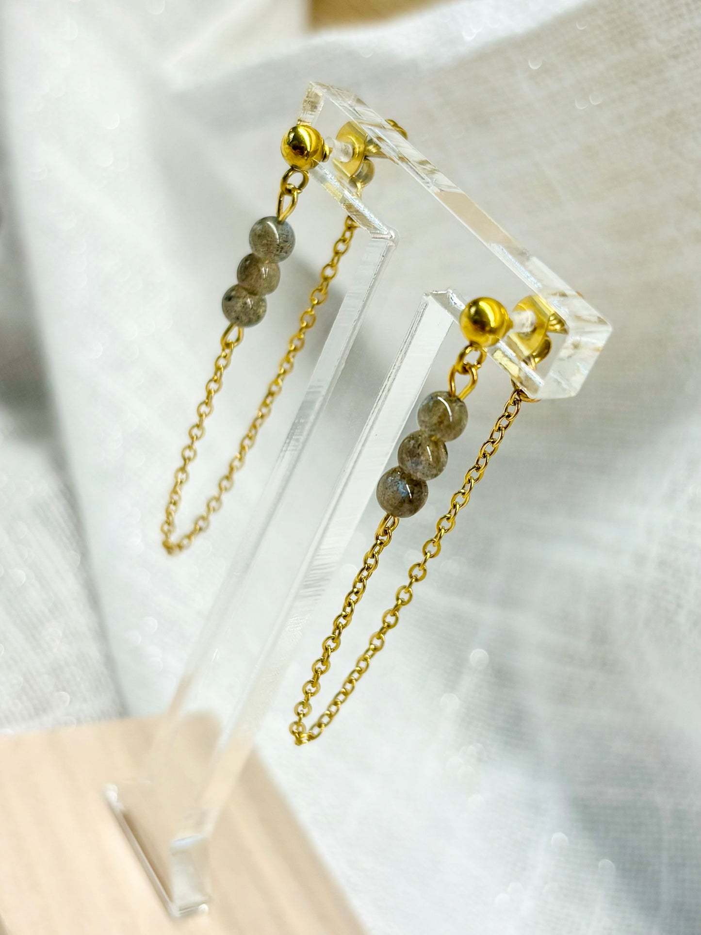Boucles d'oreille MALIA, de couleur dorée avec pierres de Labradorite, exposées sur un fond en lin.