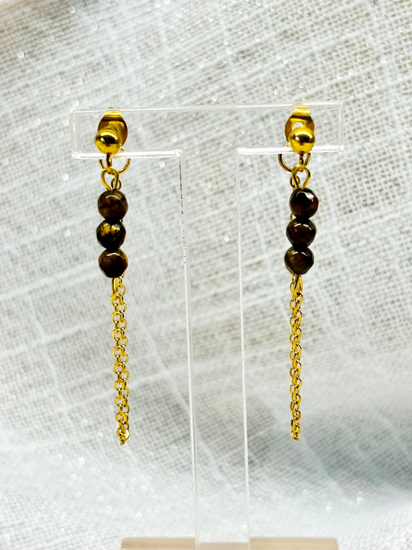 Boucles d'oreille MALIA, de couleur dorée avec pierre d'oeil de Tigre, exposées devant un fond en lin.