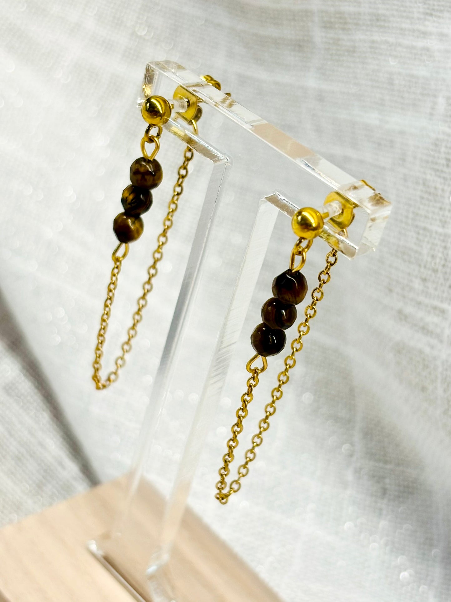 Boucles d'oreille MALIA, de couleur dorée avec pierre d'oeil de Tigre, exposées devant un fond en lin.
