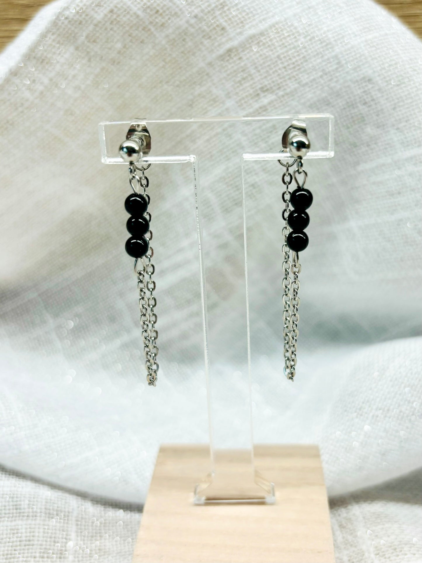 Boucles d'oreille MALIA, de couleur argent avec pierres de Tourmaline Noire, exposées devant un fond en lin.