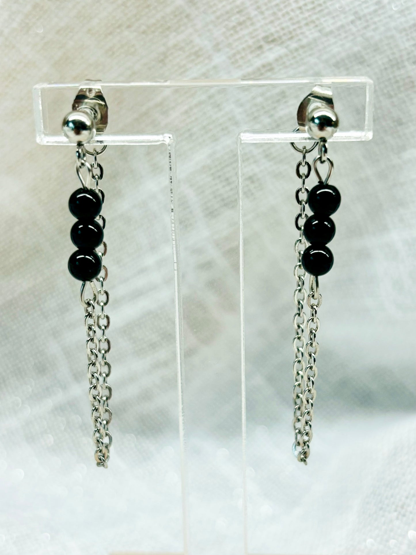 Boucles d'oreille MALIA, de couleur argent avec pierres de Tourmaline Noire, exposées devant un fond en lin.