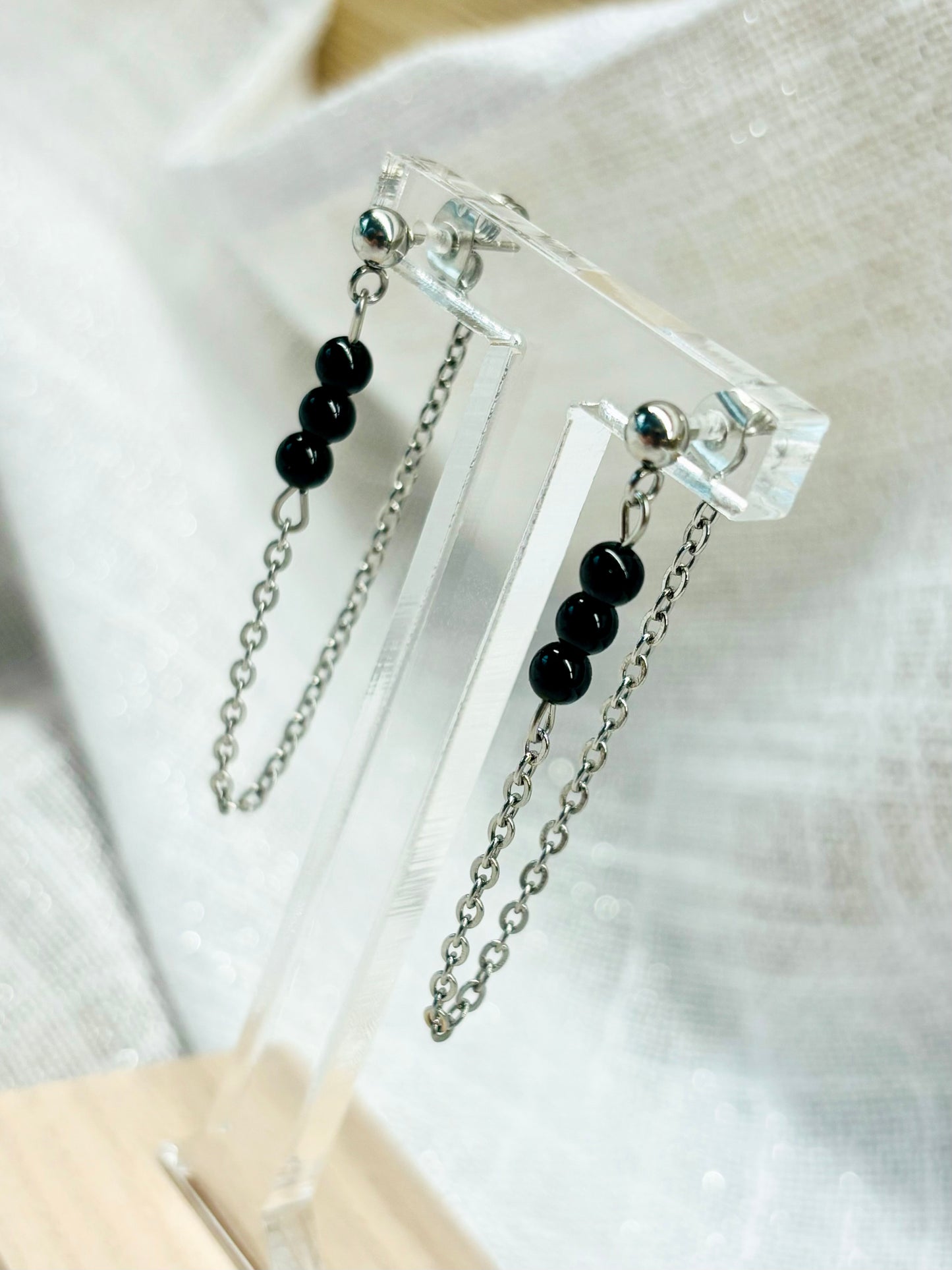 Boucles d'oreille MALIA, de couleur argent avec pierres de Tourmaline Noire, exposées devant un fond en lin.