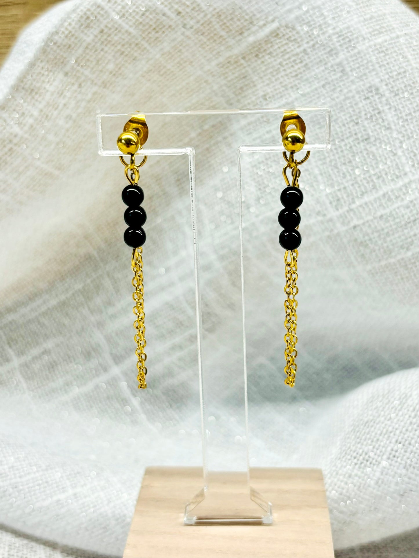 Boucles d'oreille MALIA, de couleur dorée avec pierres de Tourmaline Noire, exposées devant un fond en lin.