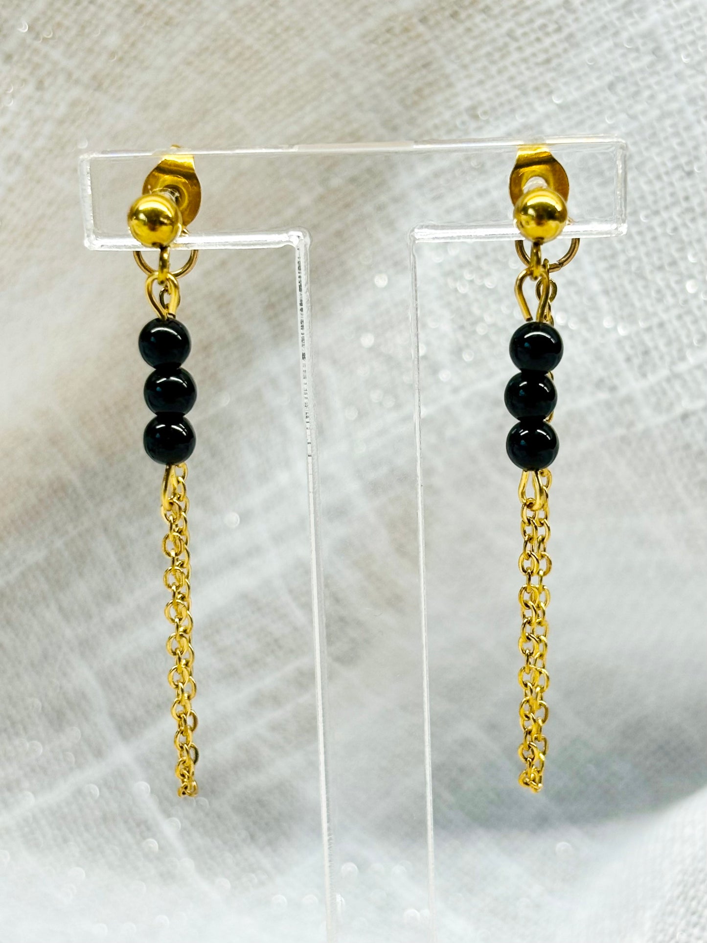 Boucles d'oreille MALIA, de couleur dorée avec pierres de Tourmaline Noire, exposées devant un fond en lin.