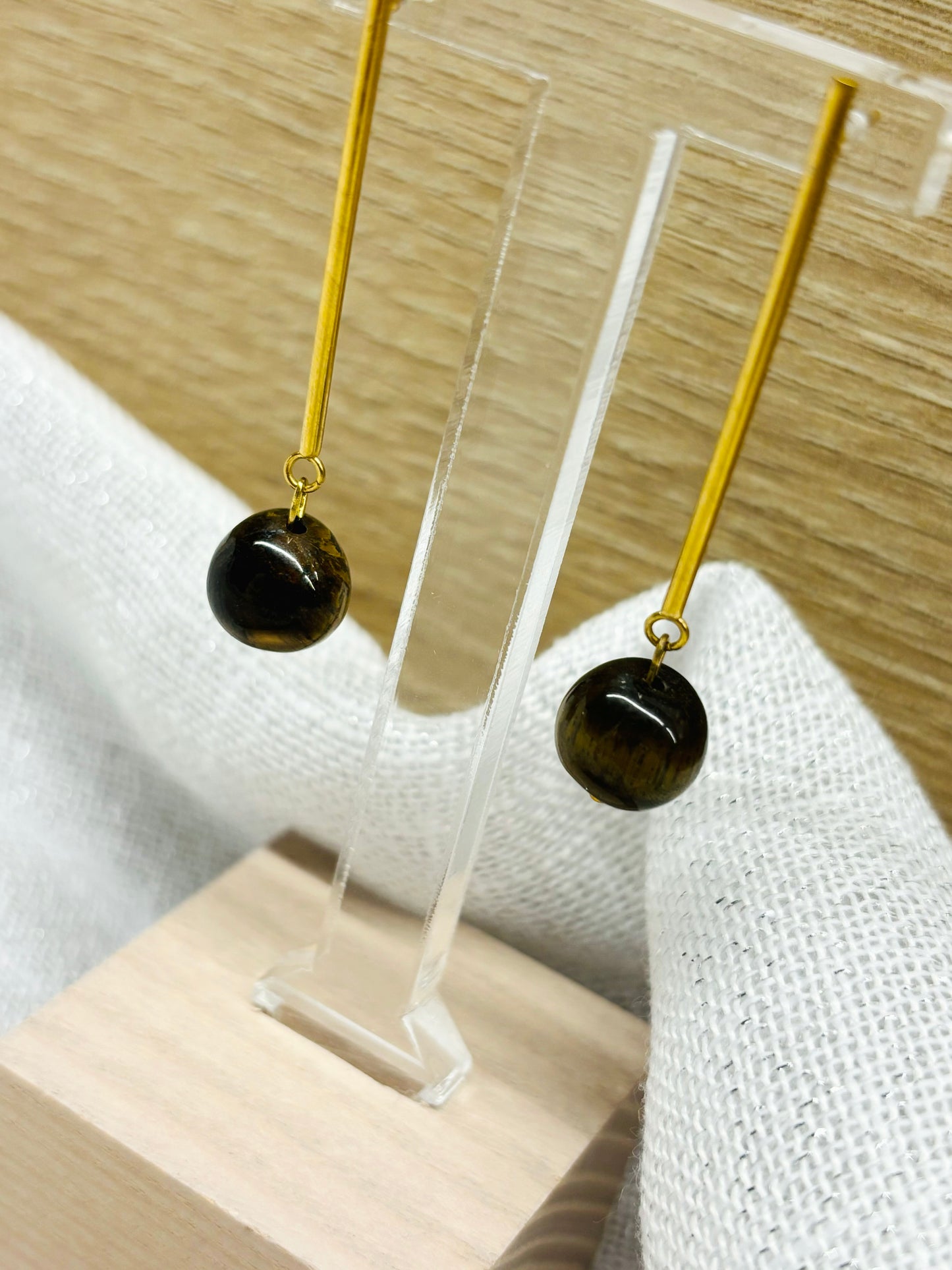 Boucles d'oreille MANON, en pierre d'Oeil de Tigre, exposées sur un fond en bois et en lin.