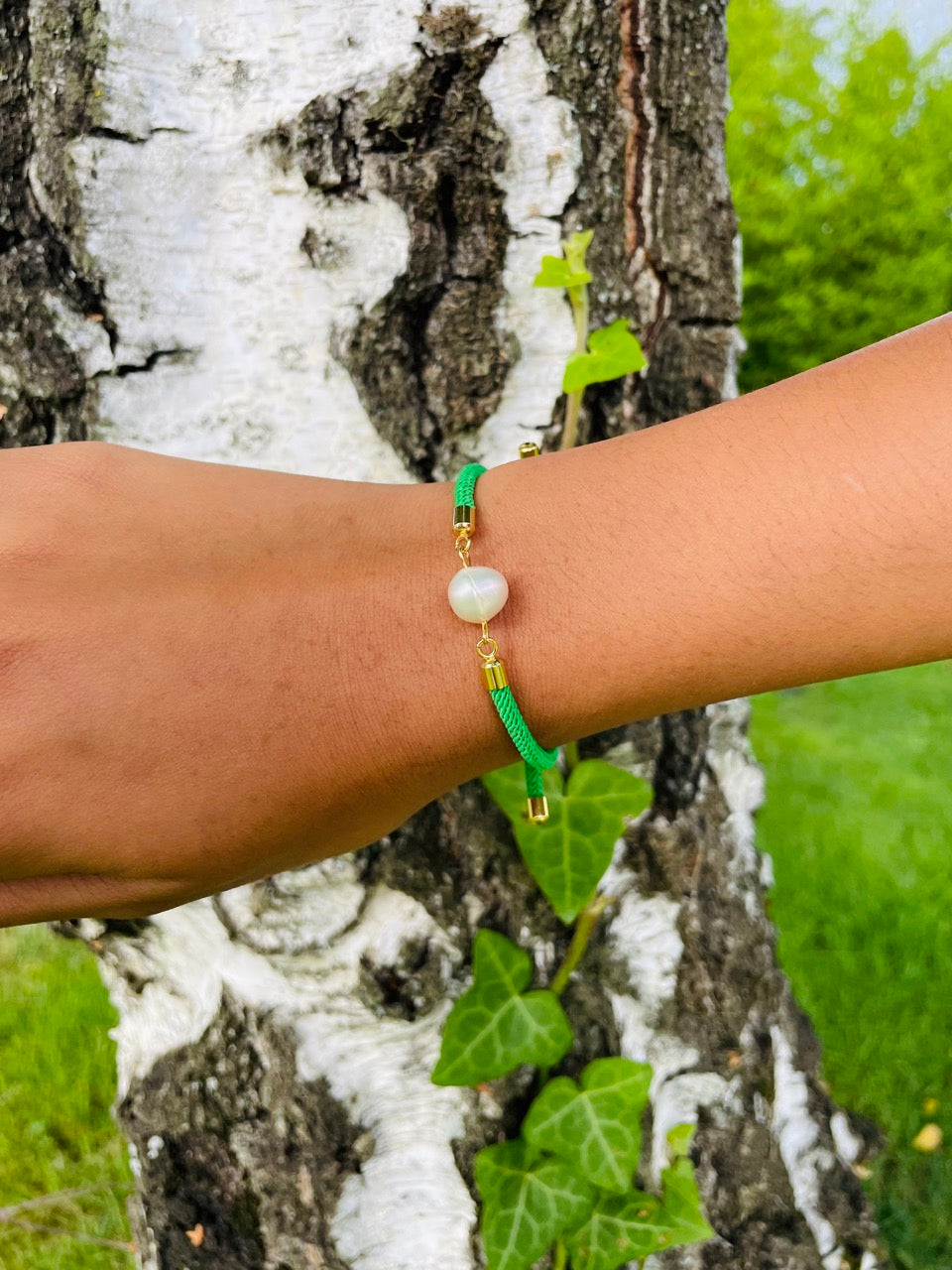 Bracelet MIA, avec cordon de couleur verte et perle de culture, porté sur un poignet de femme devant un arbre.