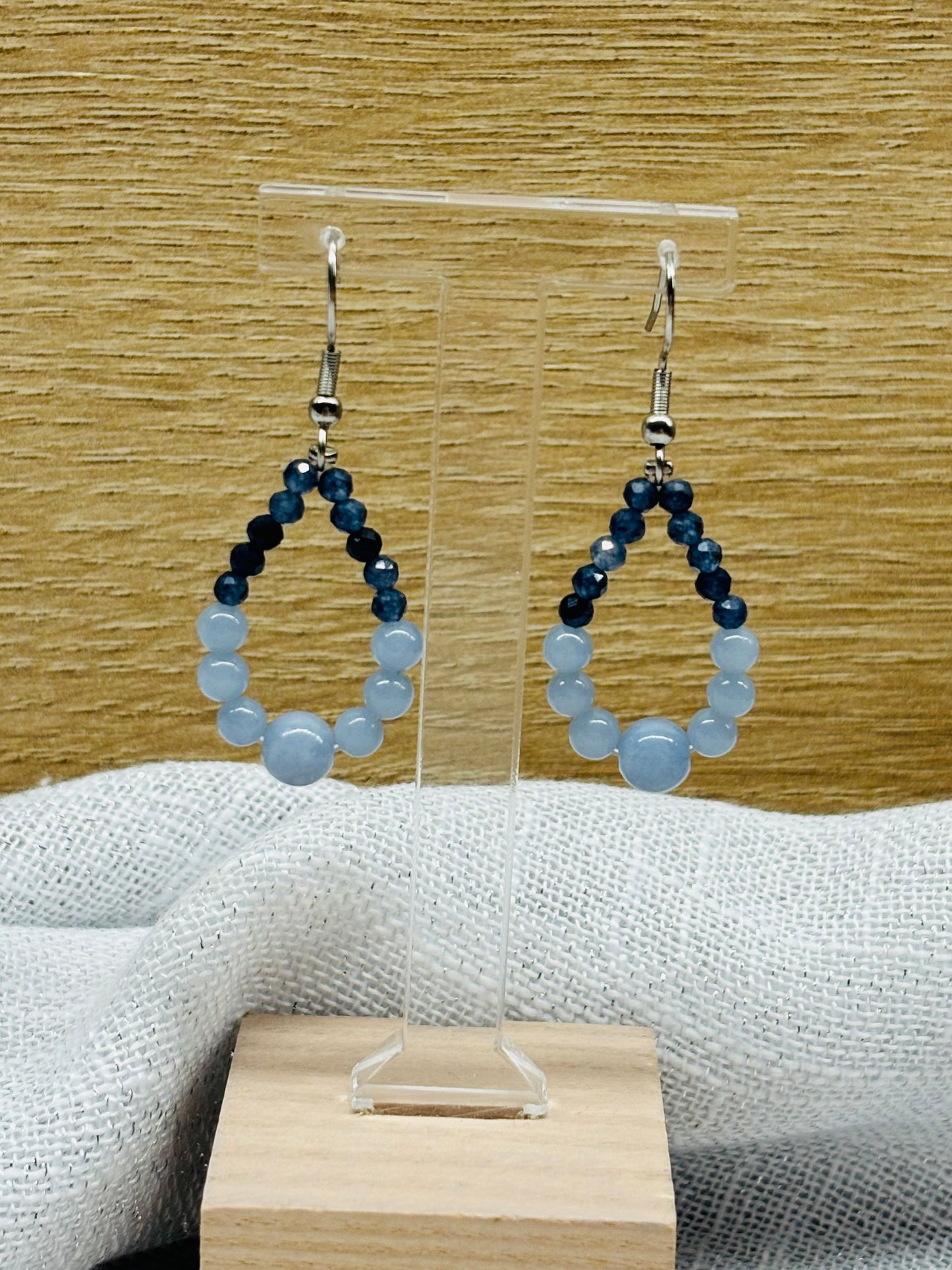 Boucles d'oreille SIMONA, avec pierres d'Angélite et de Saphir, exposées devant un fond en bois.