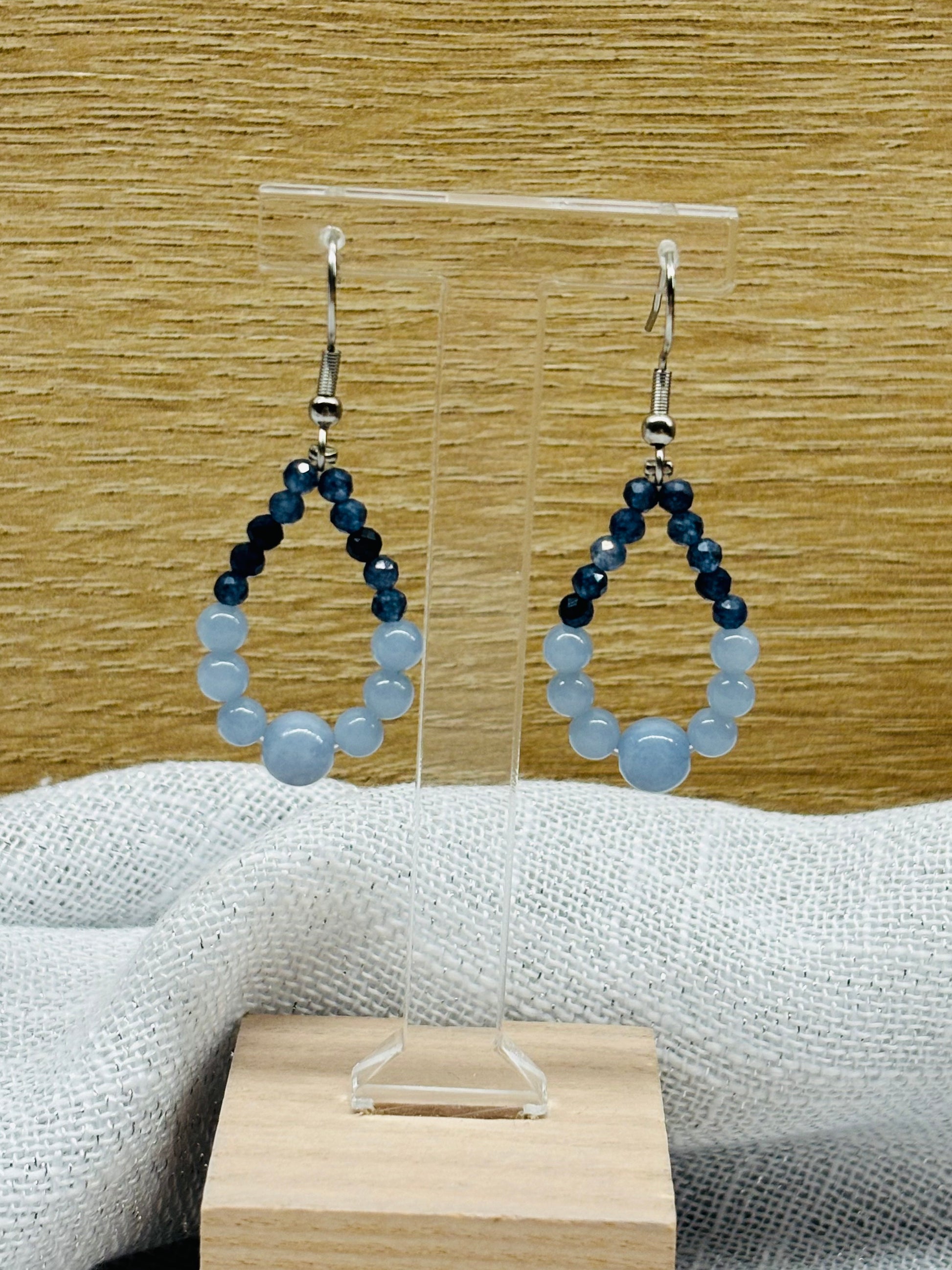 Boucles d'oreille SIMONA, avec pierres d'Angélite et de Saphir, exposées devant un fond en bois.