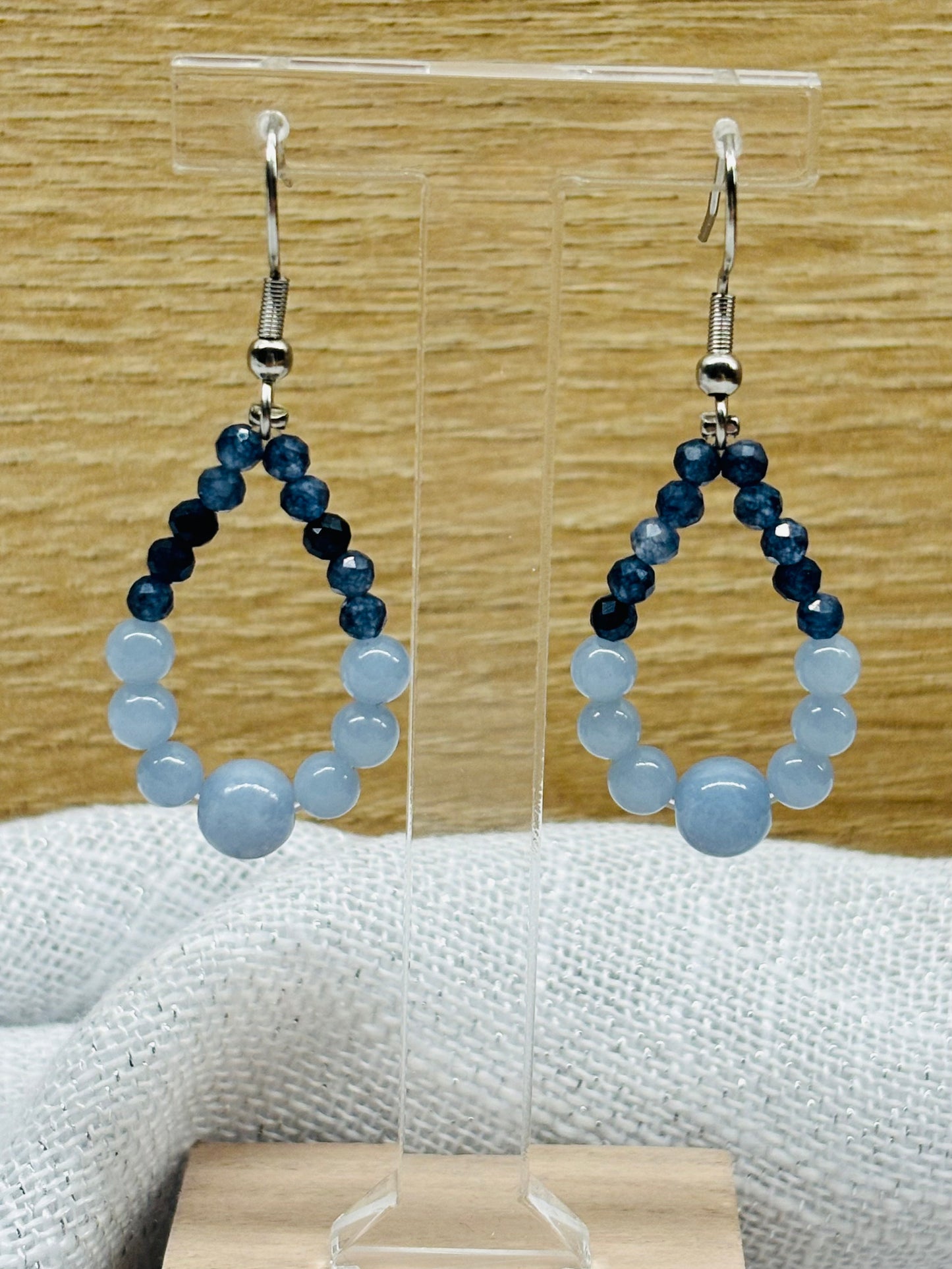 Boucles d'oreille SIMONA, avec pierres d'Angélite et de Saphir, exposées devant un fond en bois.