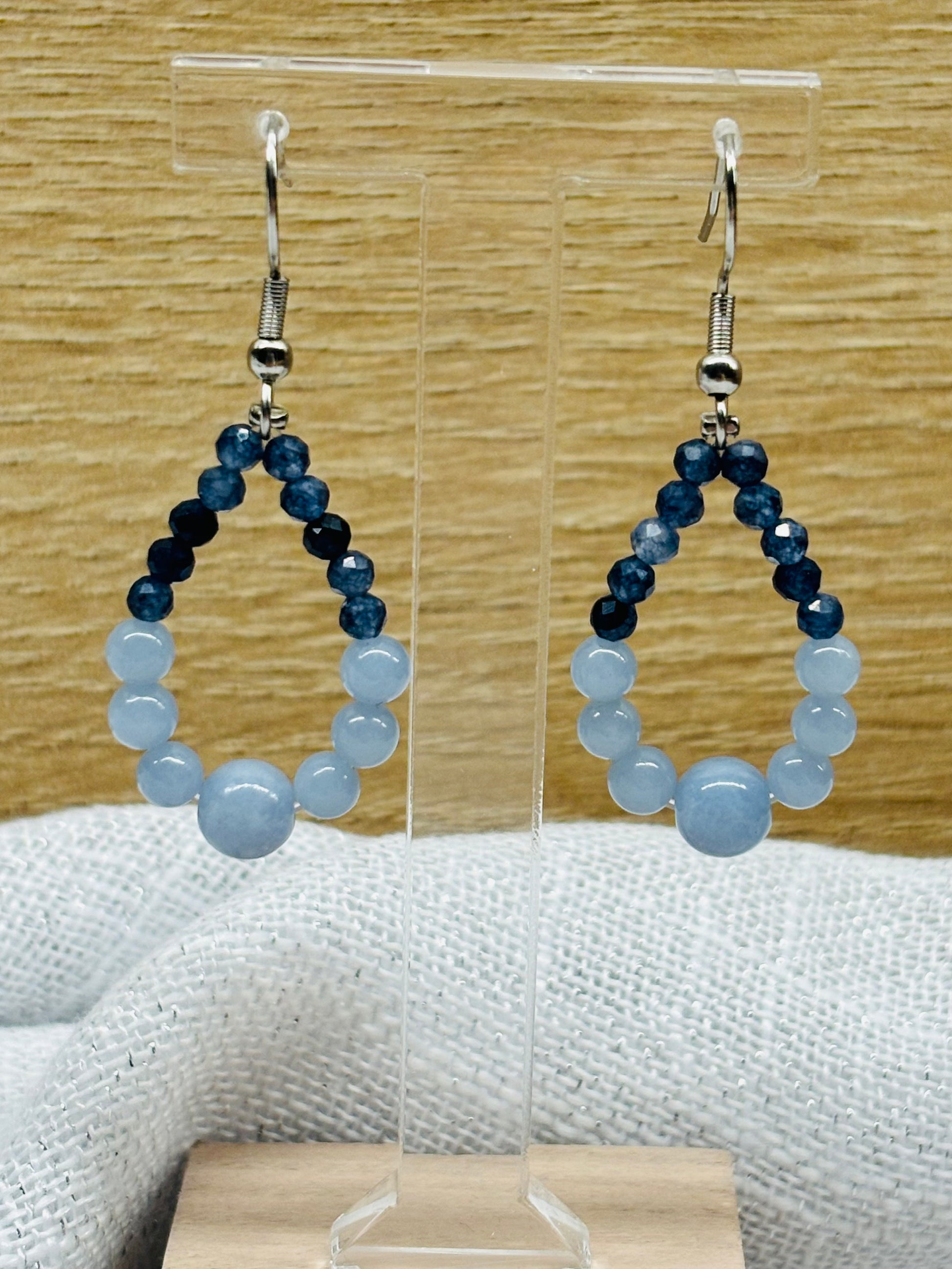 Boucles d'oreille SIMONA, avec pierres d'Angélite et de Saphir, exposées devant un fond en bois.