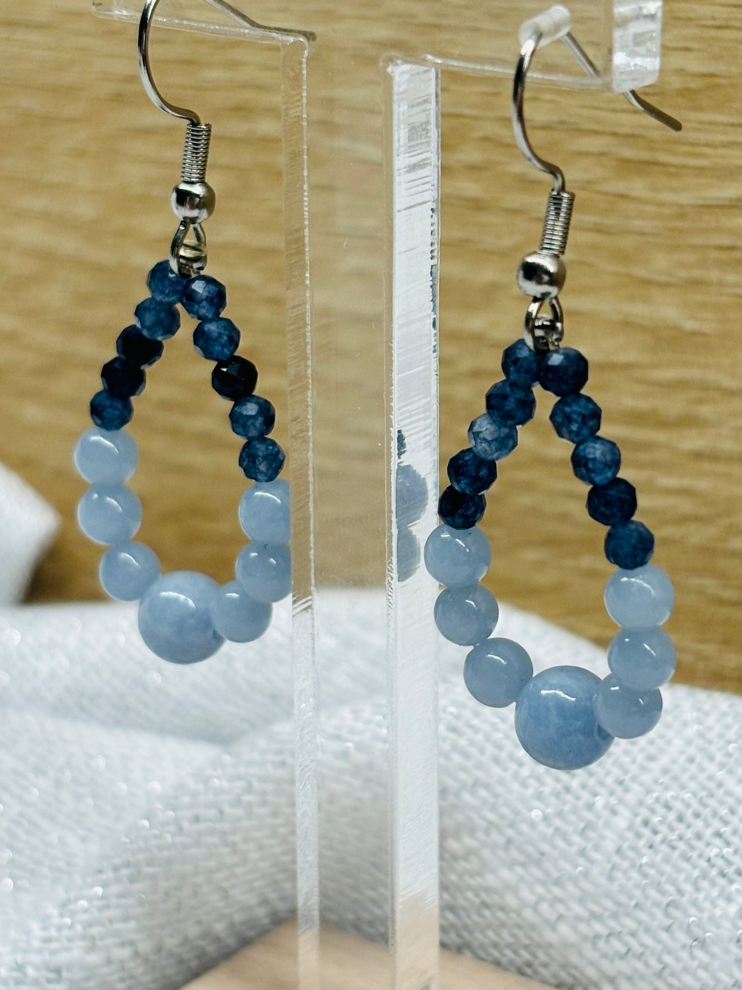 Boucles d'oreille SIMONA, avec pierres d'Angélite et de Saphir, exposées devant un fond en bois.