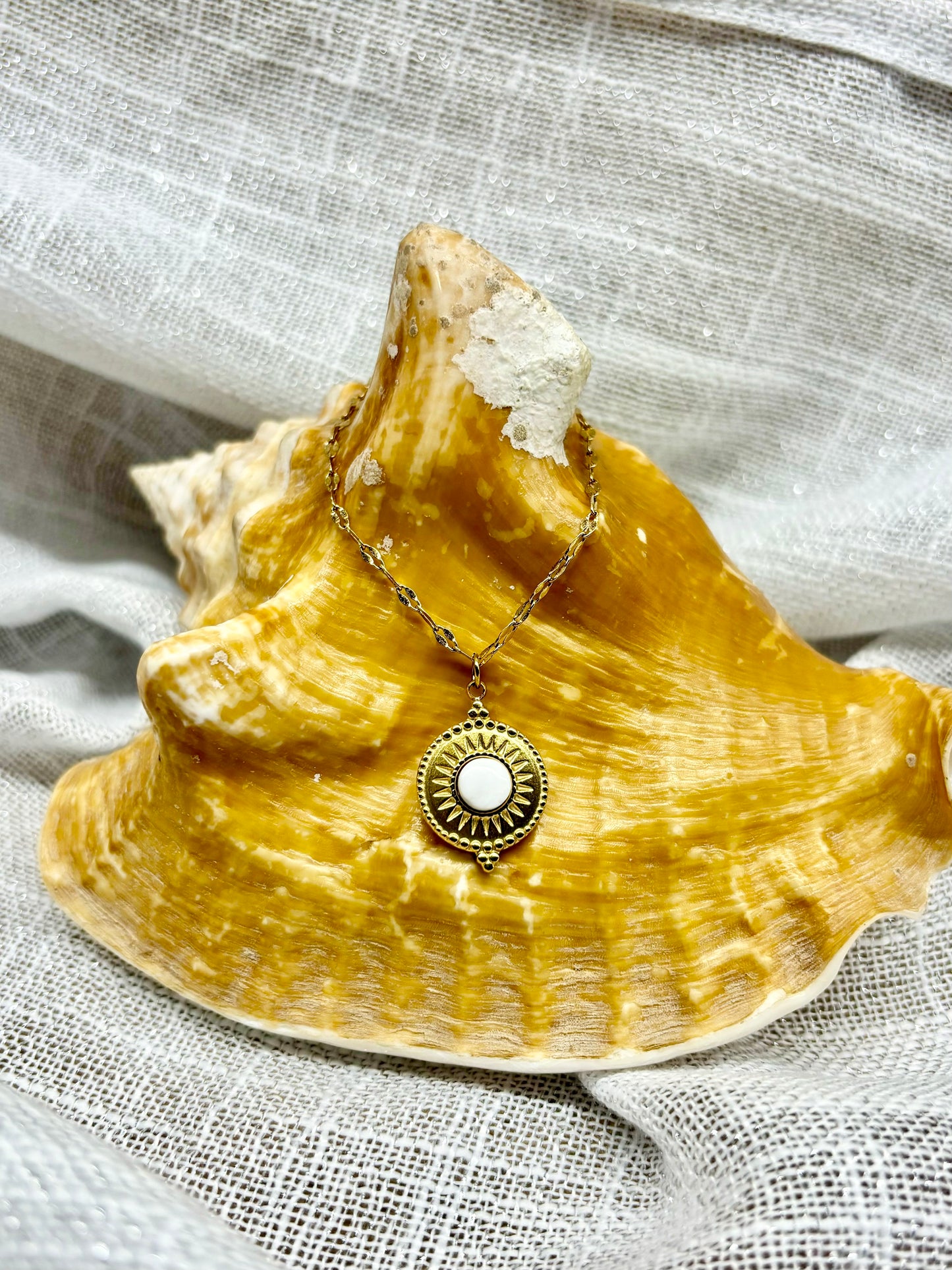 Collier ZÉLIE en acier inoxydable doré et pierre de Howlite, exposé sur un coquillage et un fond en lin.
