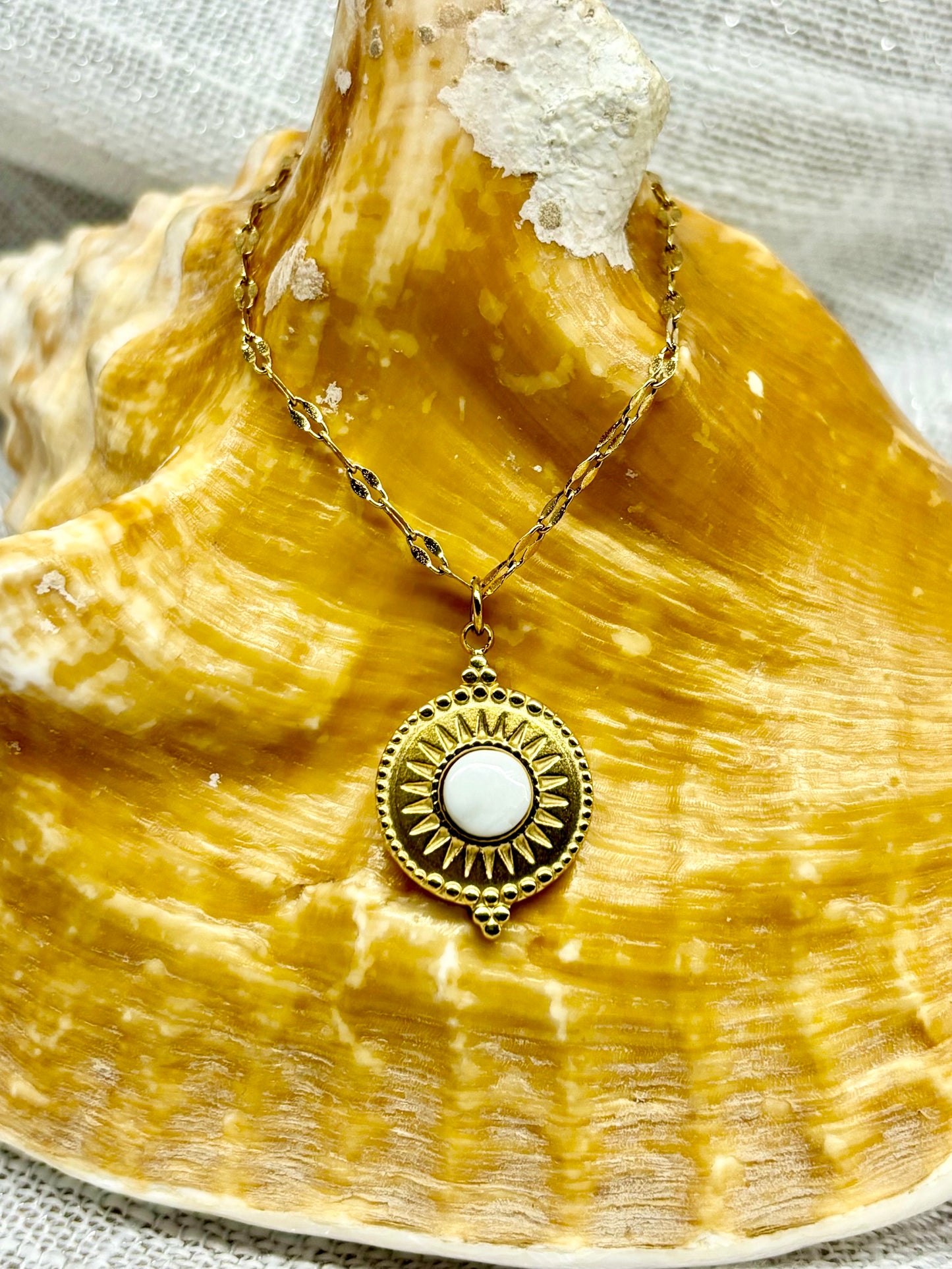 Collier ZÉLIE en acier inoxydable doré et pierre de Howlite, exposé sur un coquillage et un fond en lin.