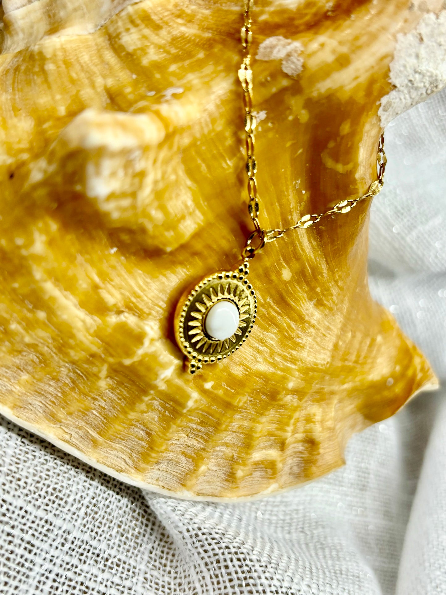Gros plan du collier ZÉLIE en acier inoxydable doré et pierre de Howlite, exposé sur un coquillage et un fond en lin.