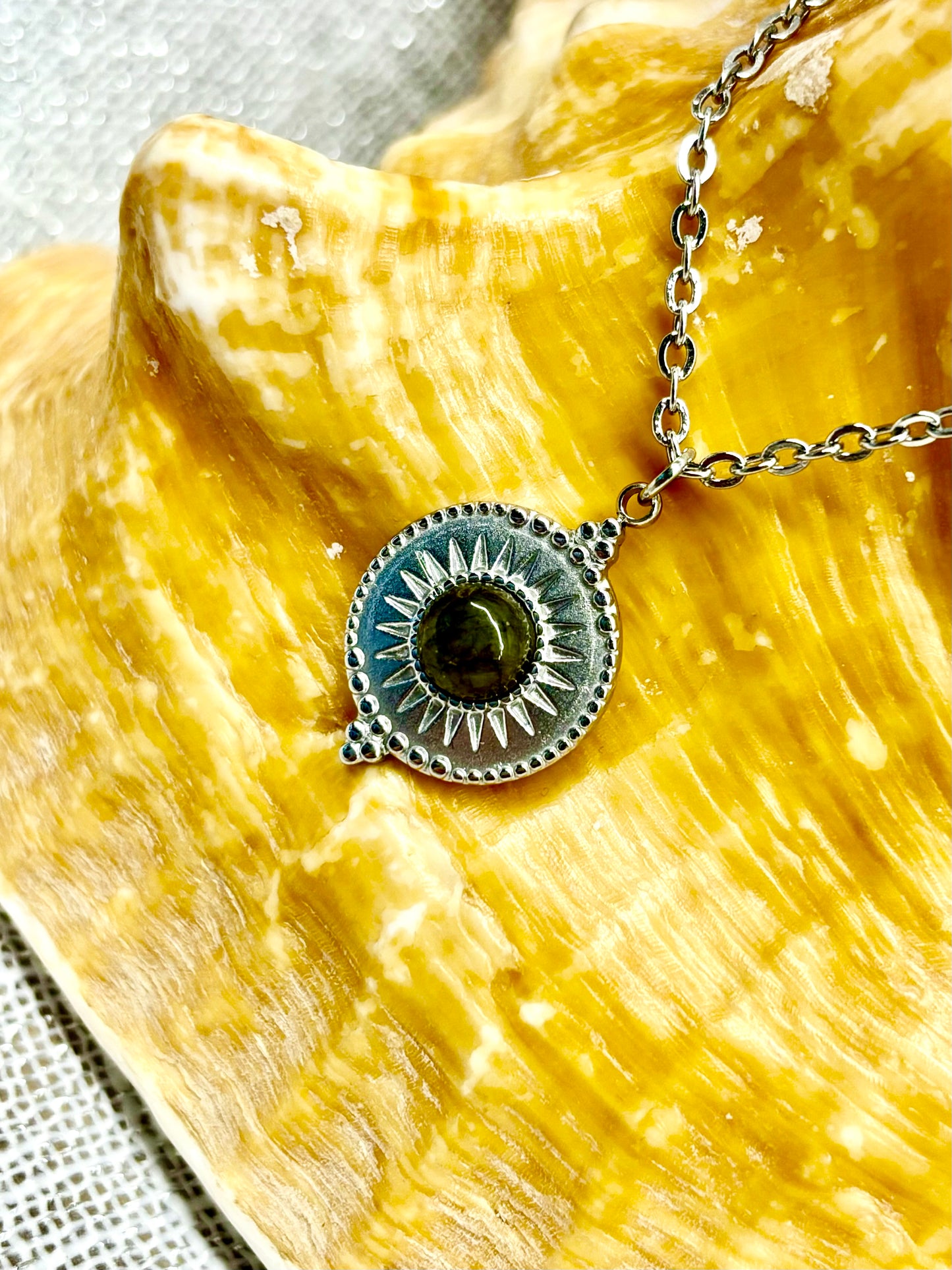 Gros plan du Collier ZÉLIE en acier inoxydable argent et pierre de Labradorite, exposé sur un coquillage et un fond en lin.