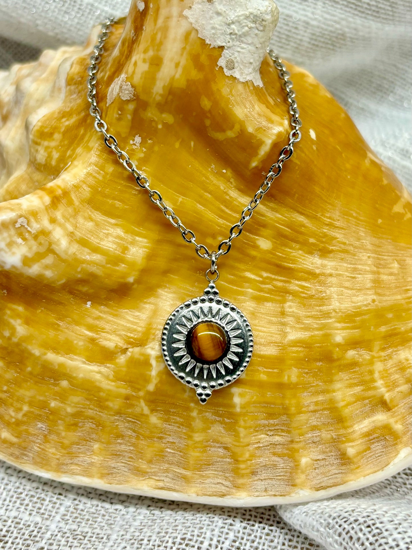 Collier ZÉLIE en acier inoxydable argent et pierre d'oeil de tigre, exposé sur un coquillage avec un fond en lin