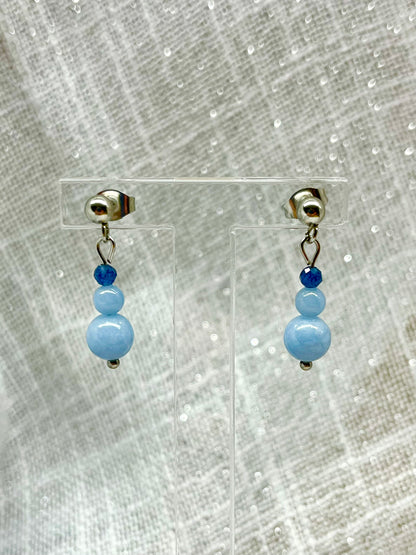 Boucles d'oreille EMMA, avec pierres d'Angélite et de Saphir, exposées devant un fond en lin.