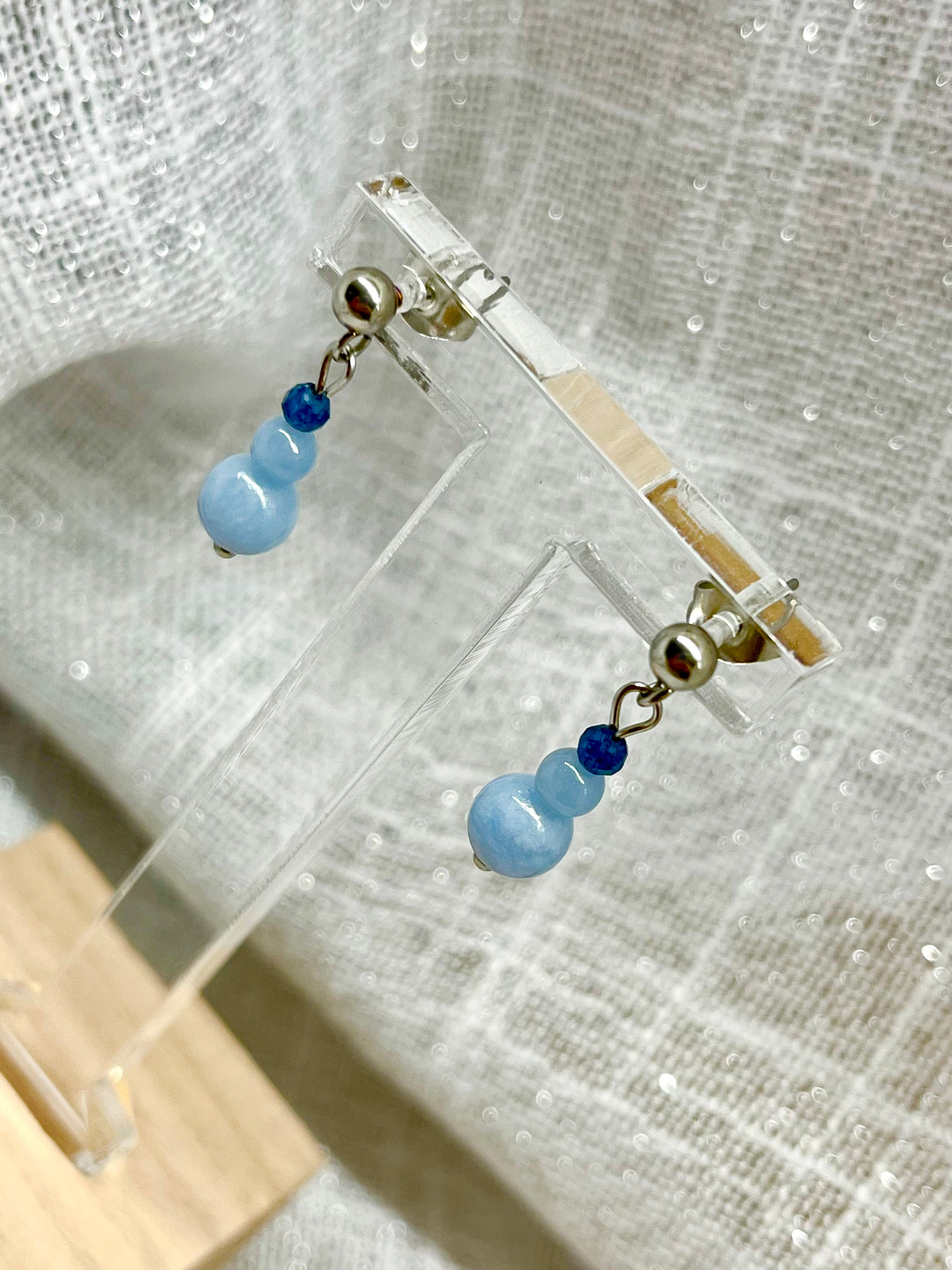 Boucles d'oreille EMMA, avec pierres d'Angélite et de Saphir, exposées devant un fond en lin.