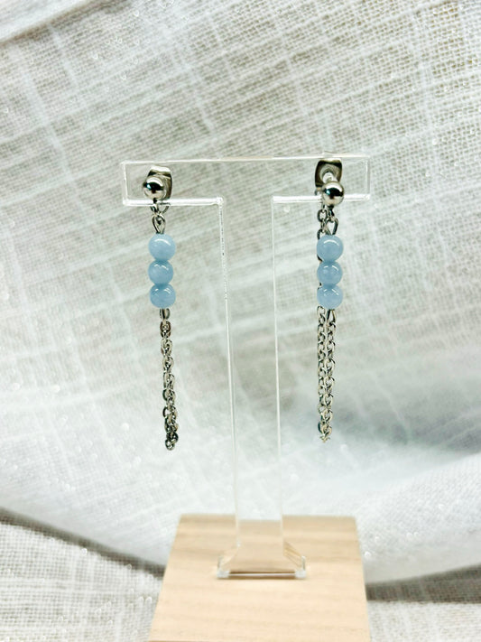 Boucles d'oreille MALIA, de couleur argent avec pierres d'Angélite, exposées devant un fond en lin.