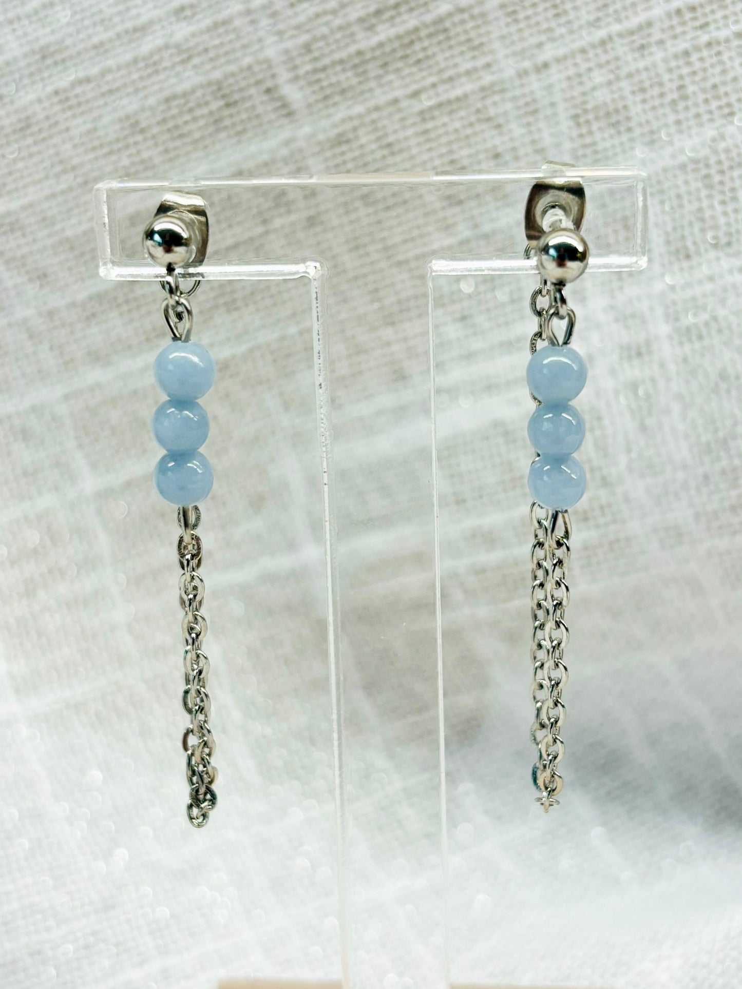 Boucles d'oreille MALIA, de couleur argent avec pierres d'Angélite, exposées devant un fond en lin.