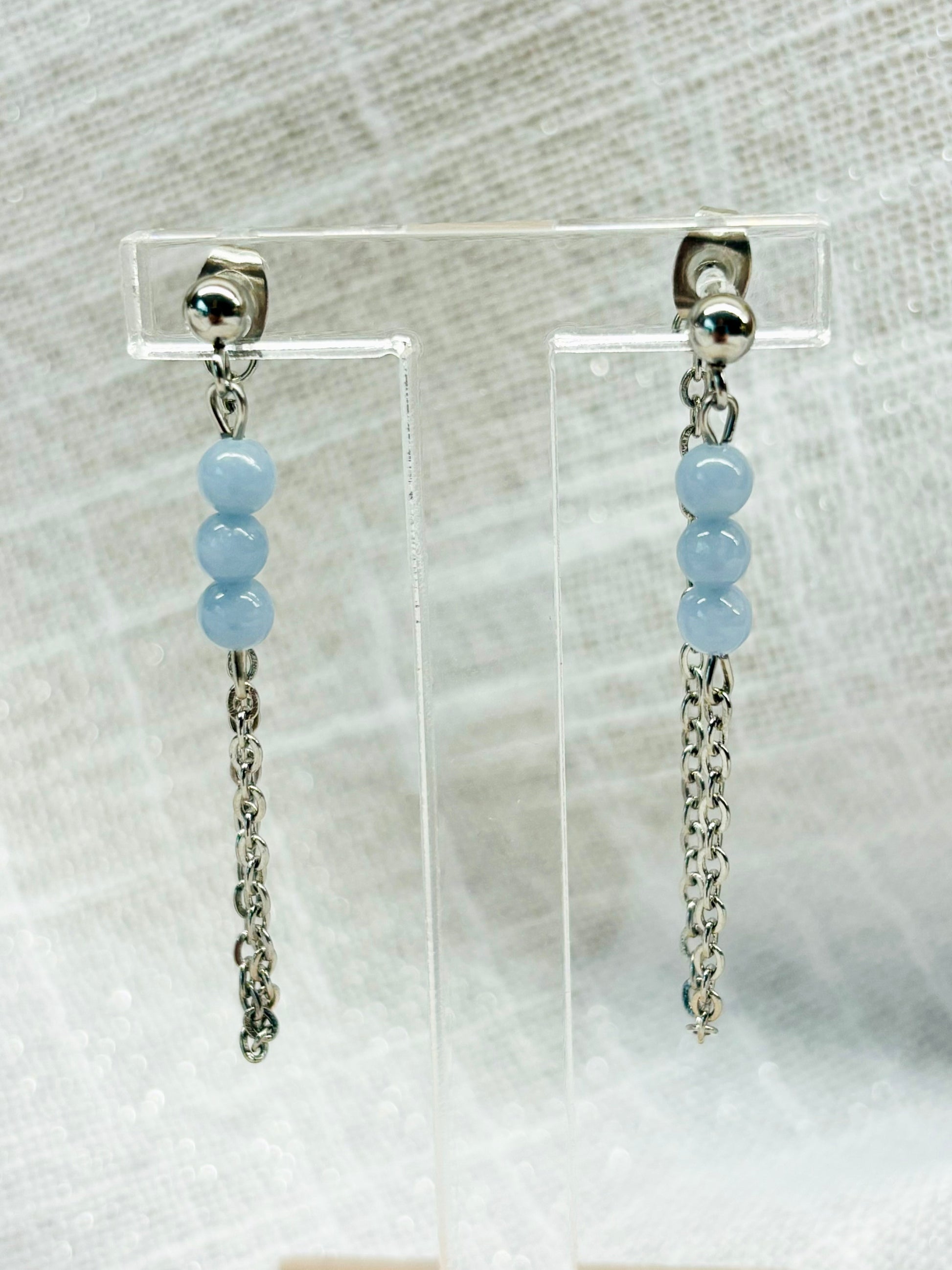 Boucles d'oreille MALIA, de couleur argent avec pierres d'Angélite, exposées devant un fond en lin.