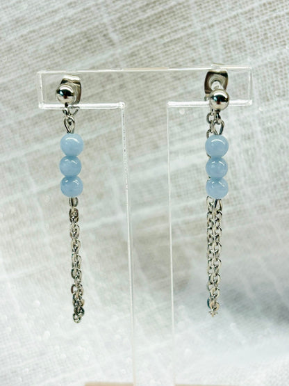 Boucles d'oreille MALIA, de couleur argent avec pierres d'Angélite, exposées devant un fond en lin.