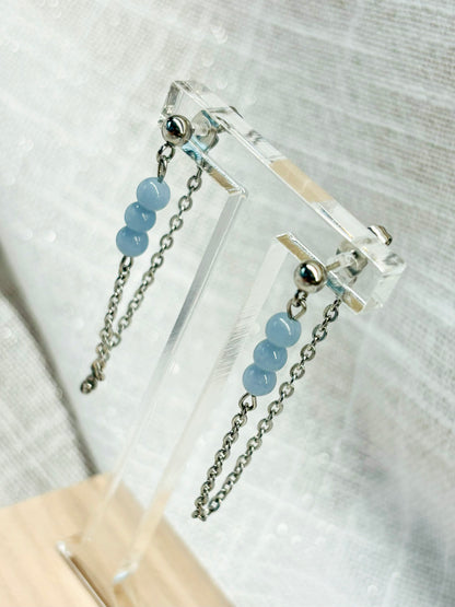 Boucles d'oreille MALIA, de couleur argent avec pierres d'Angélite, exposées devant un fond en lin.