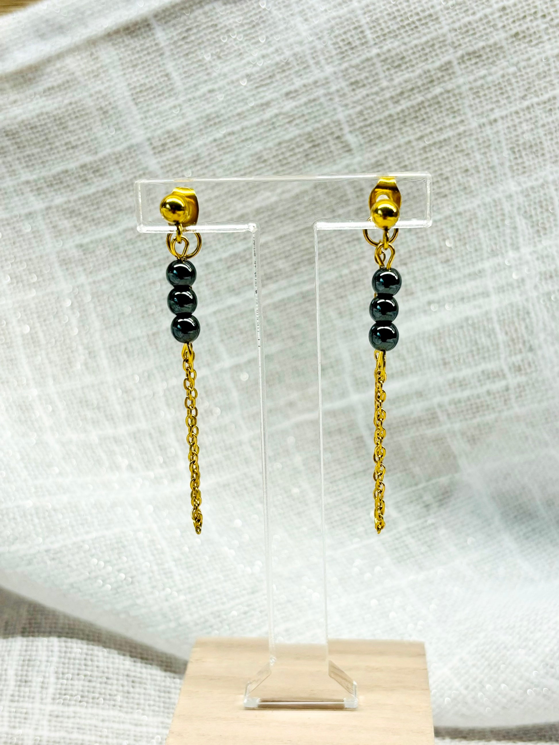 Boucles d'oreille MALIA, de couleur dorée avec pierres d'hématite, exposées sur un fond en lin.