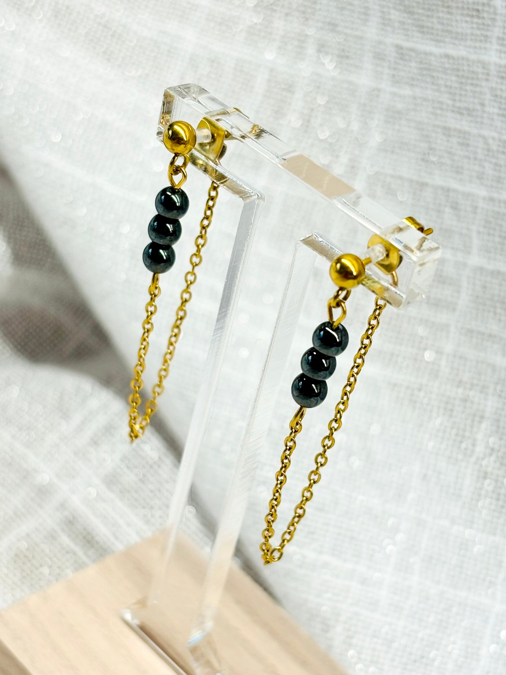 Boucles d'oreille MALIA, de couleur dorée avec pierres d'hématite, exposées sur un fond en lin.