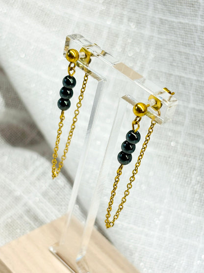 Boucles d'oreille MALIA, de couleur dorée avec pierres d'hématite, exposées sur un fond en lin.