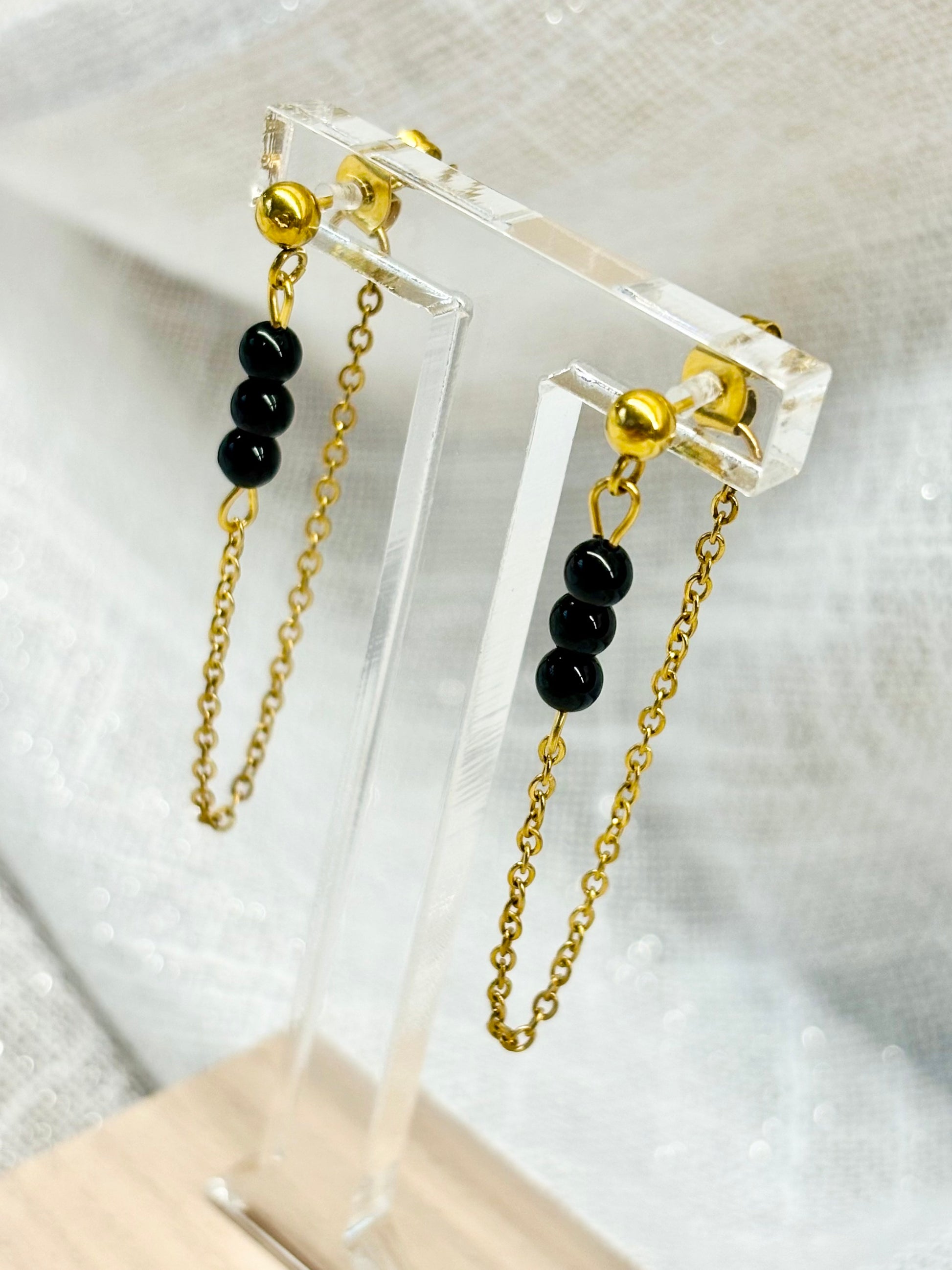 Boucles d'oreille MALIA, de couleur dorée avec pierres de Tourmaline Noire, exposées devant un fond en lin.
