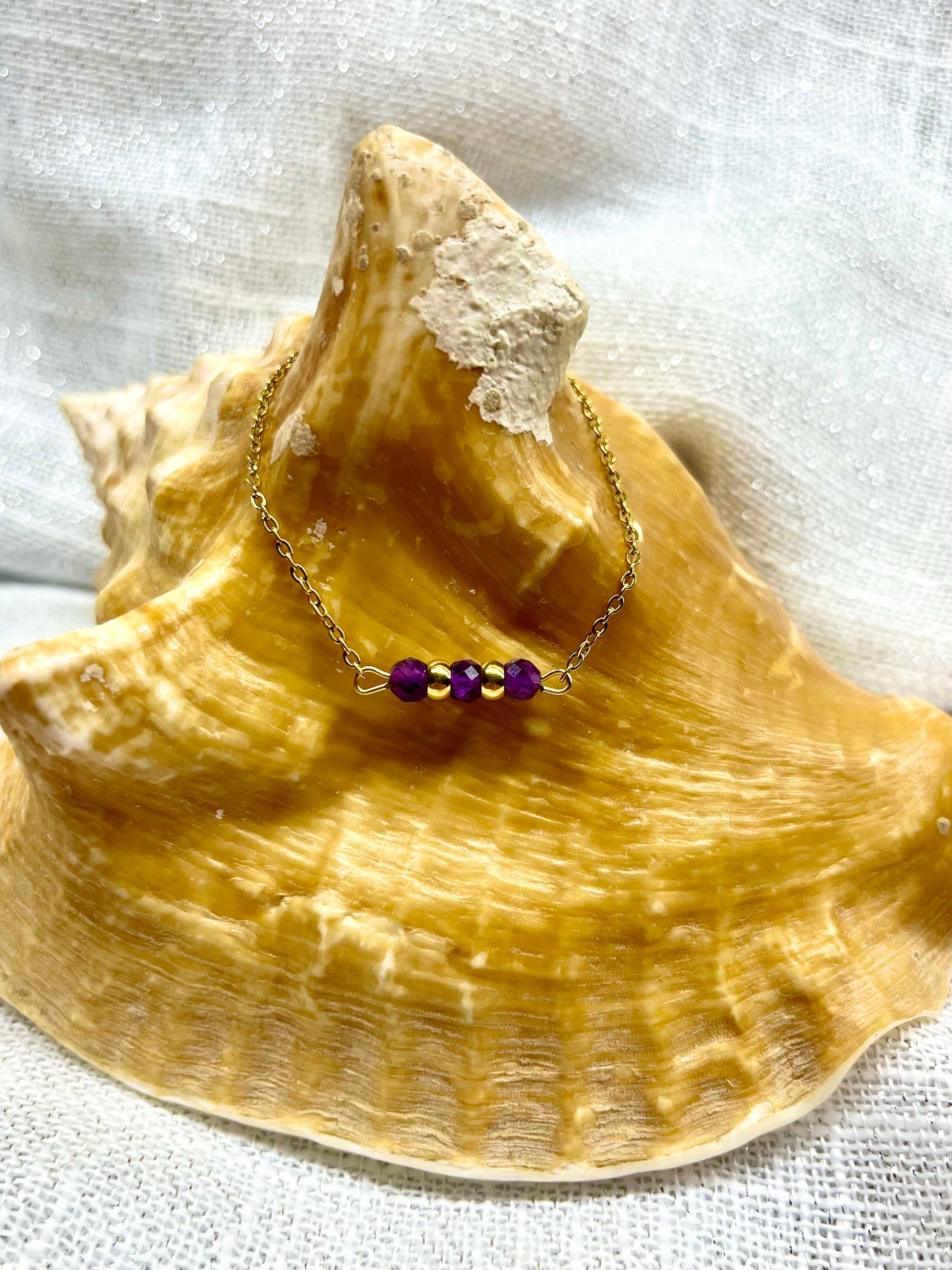 Bracelet MALIA, de couleur doré avec pierres d'Améthyste, exposé sur un coquillage.