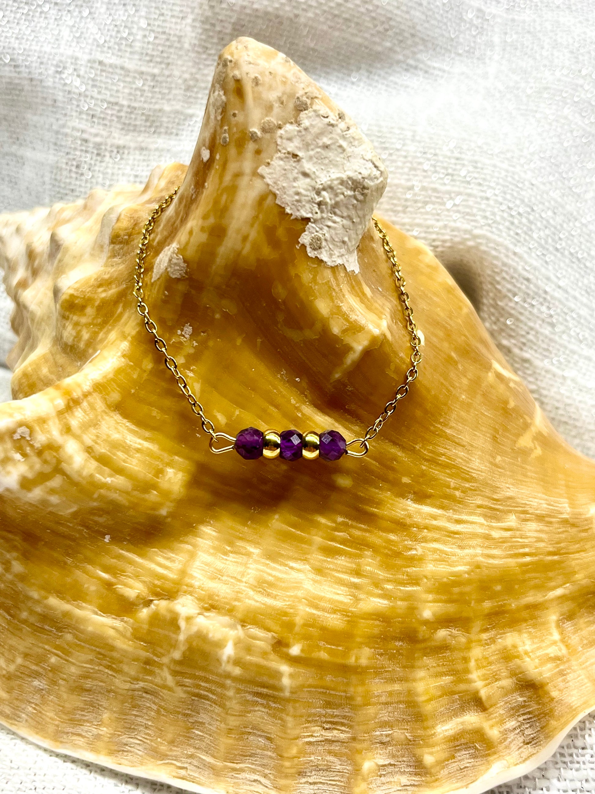 Bracelet MALIA, de couleur doré avec pierres d'Améthyste, exposé sur un coquillage.
