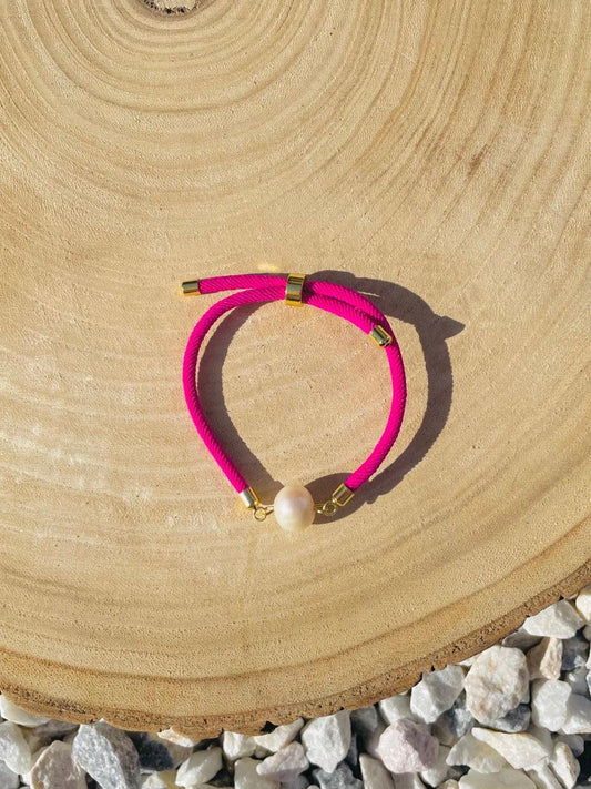 Bracelet MIA, avec cordon de couleur Fuchsia et perle de culture, exposé sur un fond en bois et graviers blancs.