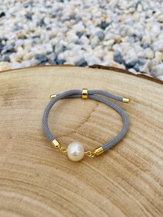 Bracelet MIA, en cordon de couleur gris souris et perle de culture, exposé sur une planche en bois et un fond de graviers blancs.