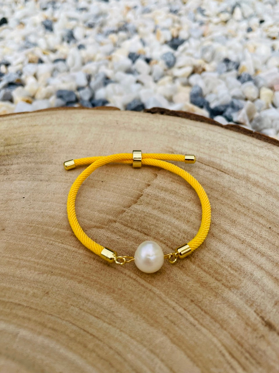 Bracelet MIA, avec cordon de couleur jaune et perle de culture, exposé sur une blanche en bois et un fond en graviers blancs.