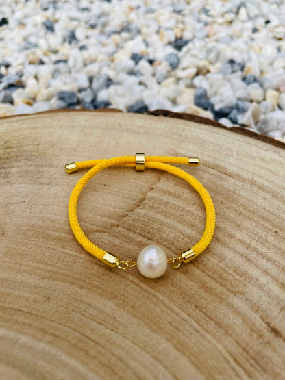 Bracelet MIA, avec cordon de couleur jaune et perle de culture, exposé sur une blanche en bois et un fond en graviers blancs.