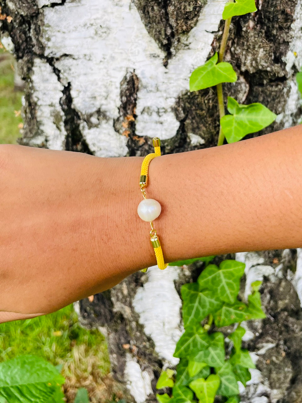 Bracelet MIA, avec cordon de couleur jaune et perle de culture, porté sur un poignet de femme devant un arbre.