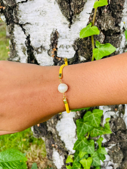 Bracelet MIA, avec cordon de couleur jaune et perle de culture, porté sur un poignet de femme devant un arbre.