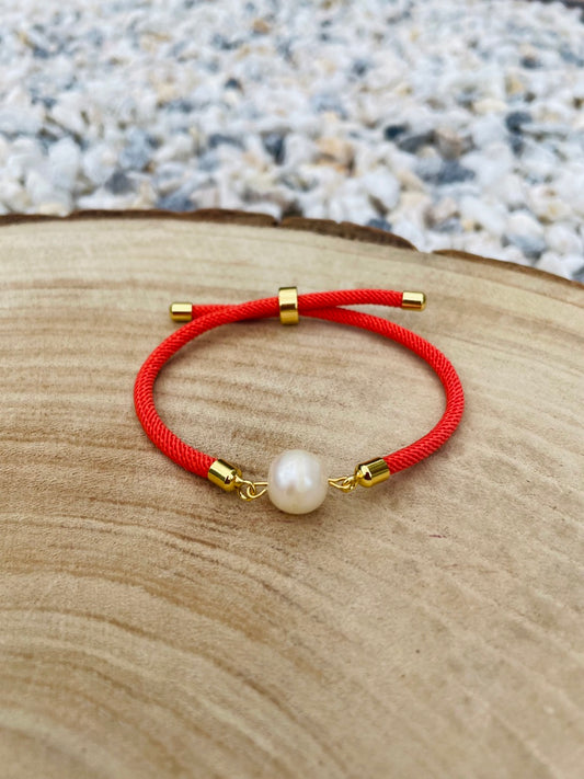 Bracelet MIA, avec cordon de couleur orange et perle de culture, exposé sur un fond en bois et graviers blancs.
