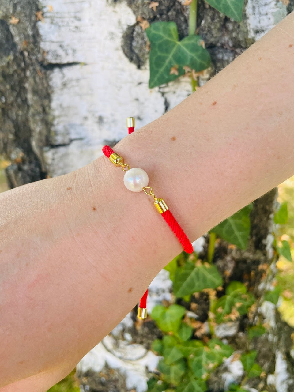 Bracelet MIA, avec cordon de couleur rouge et perle de culture, porté sur un poignet de femme devant un arbree.