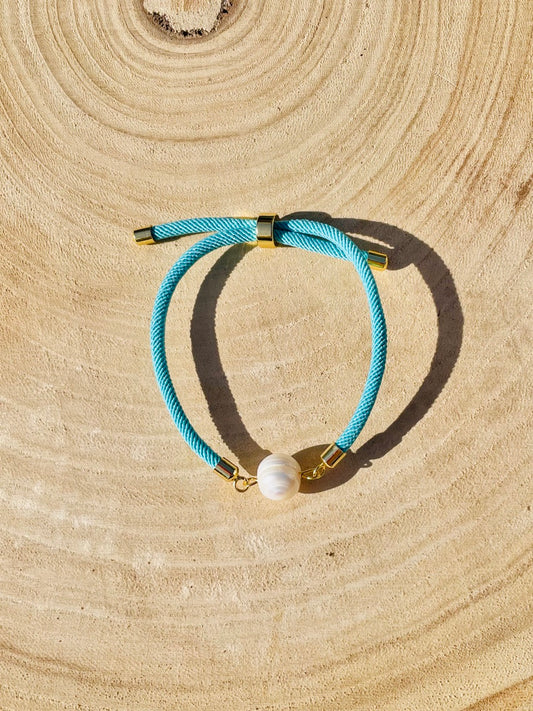 Bracelet MIA, avec cordon de couleur turquoise et perle de culture, exposé sur une planche en bois et un fond de graviers blancs.