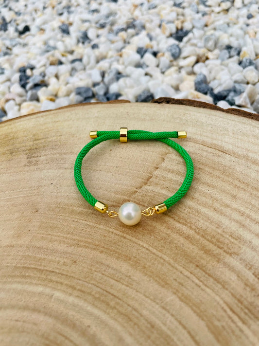 Bracelet MIA, avec cordon de couleur verte et perle de culture, exposé sur un fond en bois et graviers blancs.