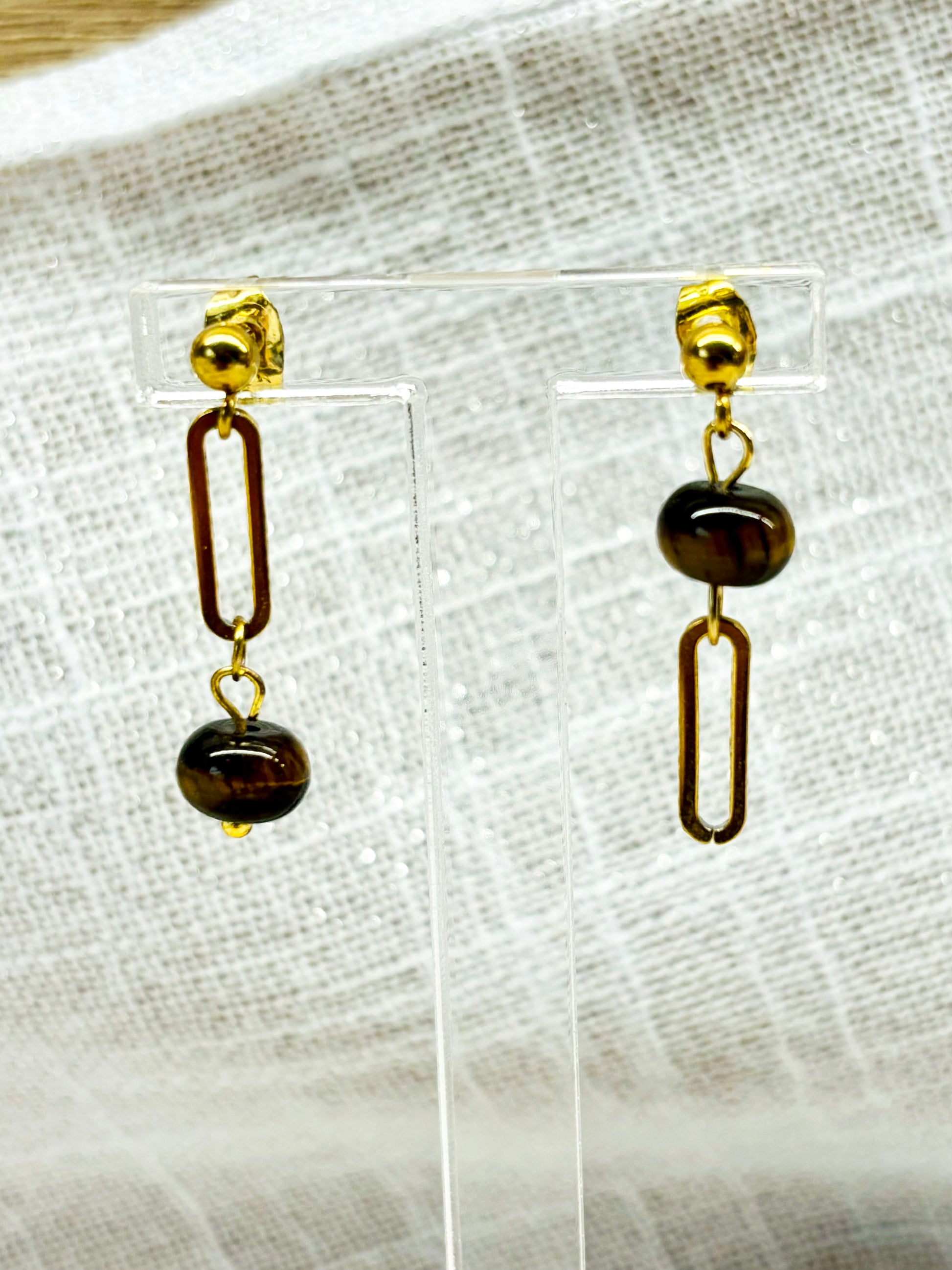 Boucles d'oreille NOËLA, avec pierre d'Oeil de Tigre, exposées sur un fond en lin.